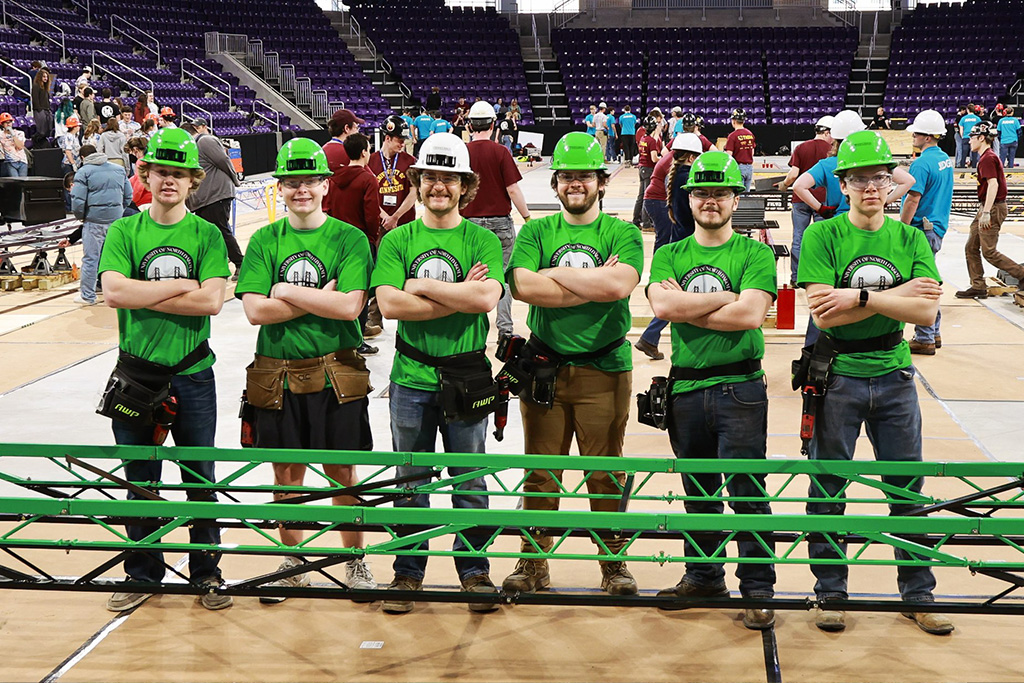 A group of students wearing hard hats and green shirts stand with crossed arms for a group photo behind a constructed steel bridge inside of a gymnasium.