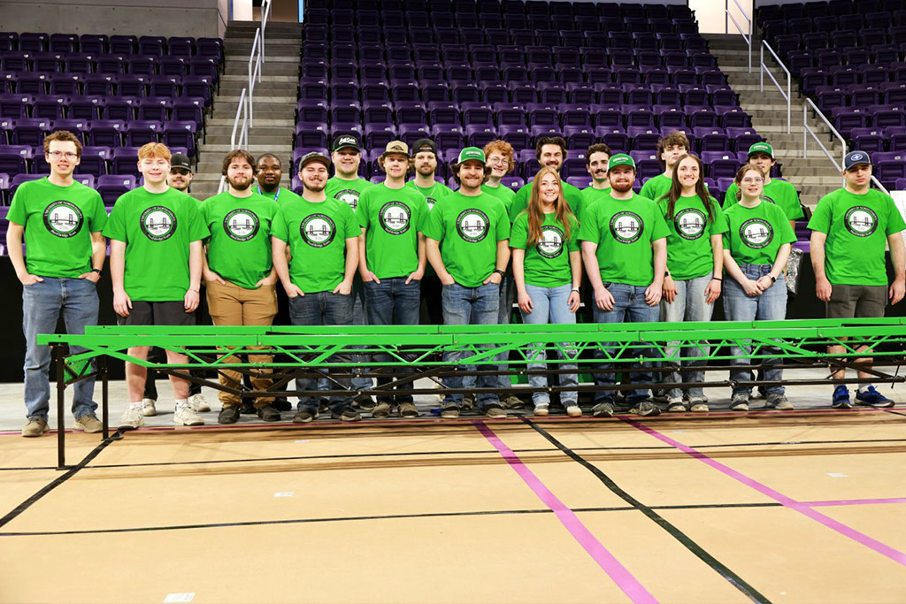 A large group of students wearing UND green shirts stand in front of a bright green steel bridge constructed inside of a gymnasium.