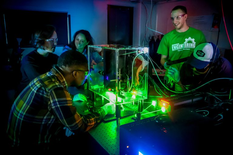 Students and a faculty member work inside of a dark lab that is illuminated by green and blue LED equipment.