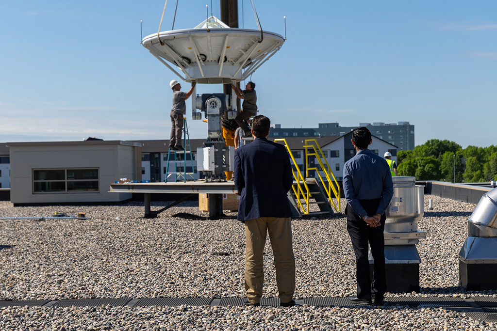 Two people stand and watch as two other people in hardhats work on an antenna being installed on a rooftop.