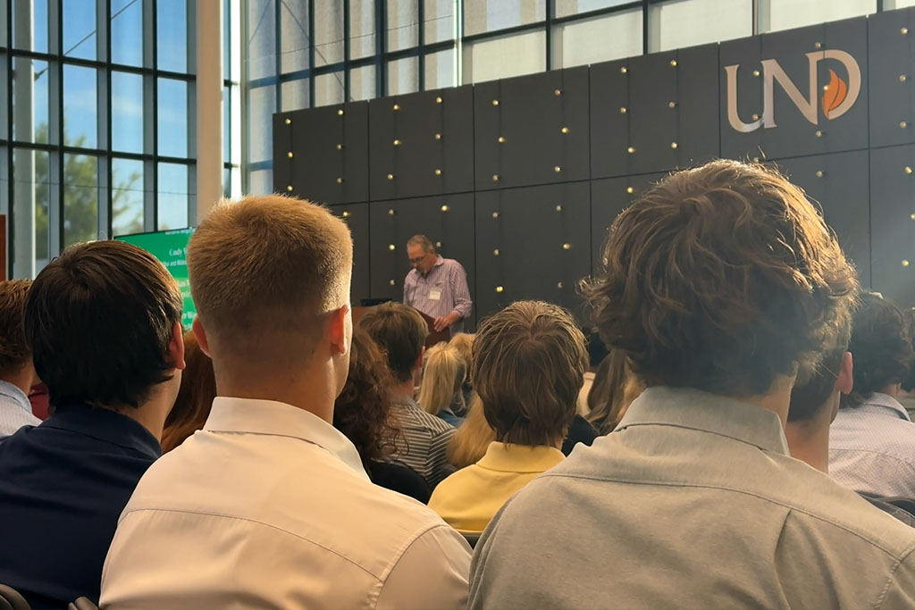 In a formal ceremony, students listen to a speaker at a podium inside of a brightly lit room.