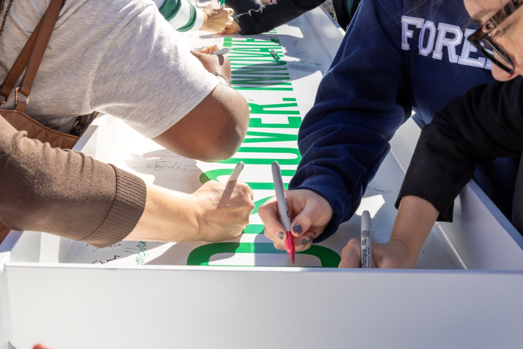 Several people lean over to sign their names in Sharpie on a white steel beam.