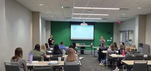 Two presenters stand at the front of a classroom in front of students. A large projector is in the background.