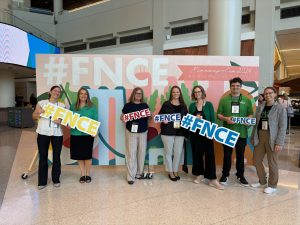 N&D Faculty & staff hold signs with marketing slogans in front of a photo backdrop at the FNCE conference.