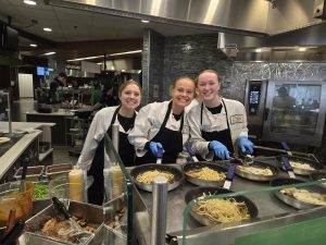 Three students stand behind a food warmer, with pasta in pans prepared to serve.