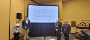 Two students with two faculty stand in front of a projector display.