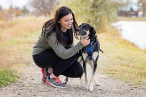 McKenzie Moe kneels down next to her dog. She is on a dirt parth with grass on either side.