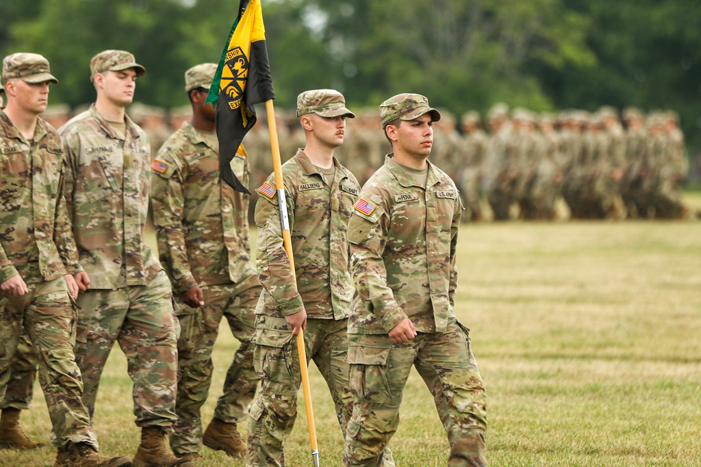 ROTC cadets in uniforms march across a field. Cadet Hallberg holds a flag on a long poll. The flag is yellolack and features an ROTC logo.