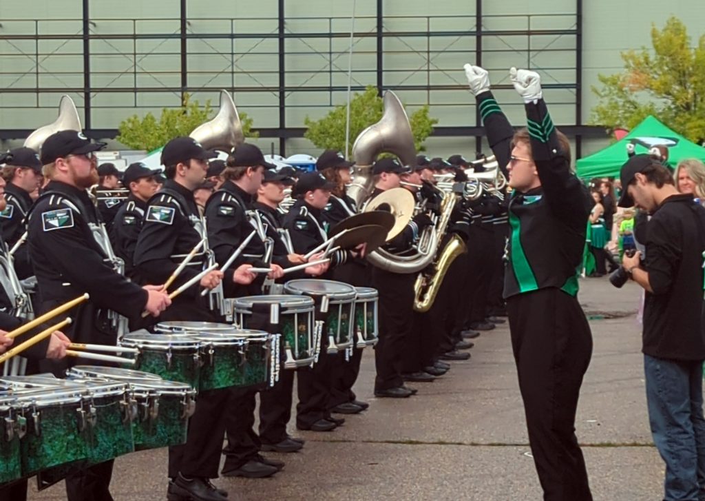 A band director lifts his arms in the air in front of a row of drummers.