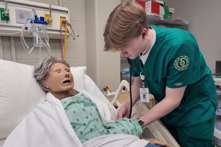 A nursing student in green scrubs leans over a patient simulator maniken in a hospital bed.