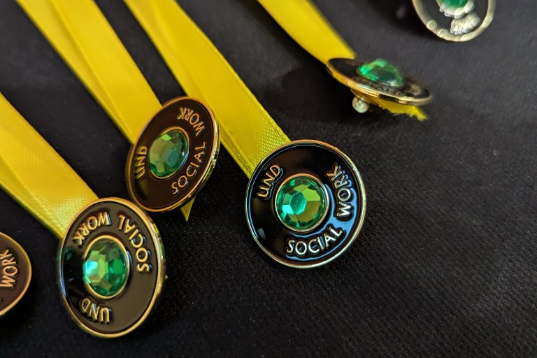 A row of UND Social Work pins sit on a table with yellow ribbon running behind.