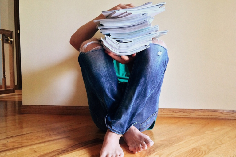 Person sitting on a wooden floor, knees up. They are holding a messy stack of journals on their knees.