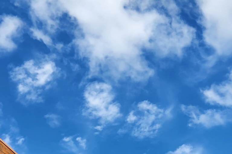 This is a picture of a bright blue sky filled with whispy white clouds. In the bottom left corner there is a piece of wood that crosses the picture.