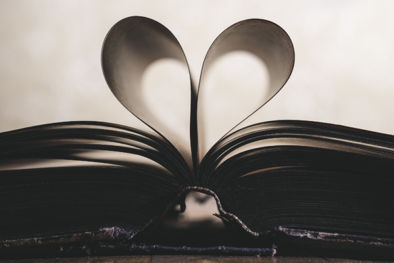 A book lays open on a wooden table. The center two pages are folded in on themselves to form a heart. The photo has an ivory background and the book is in the shade so the pages are all either black or dark gray.