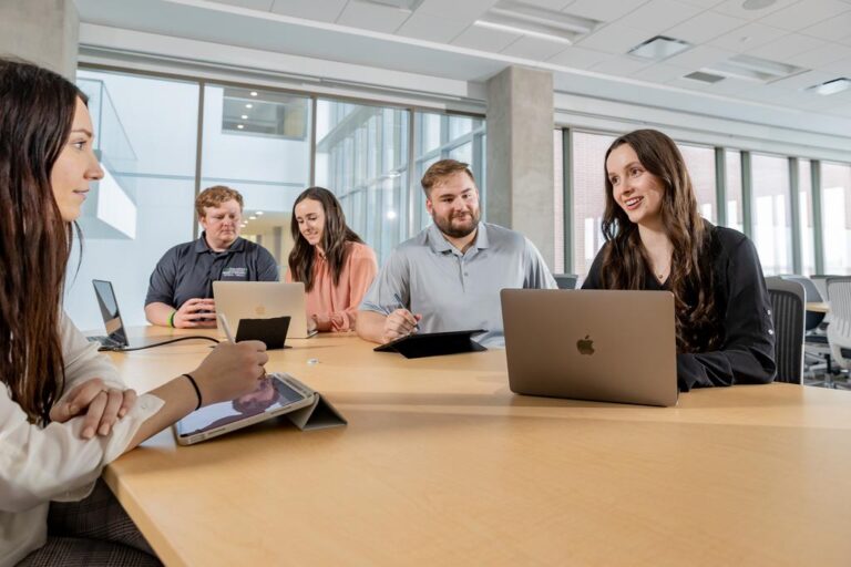 People are sitting around a table, some looking at laptops and some looking at tablets. They appear to be in smaller groups, conversing in those groups.