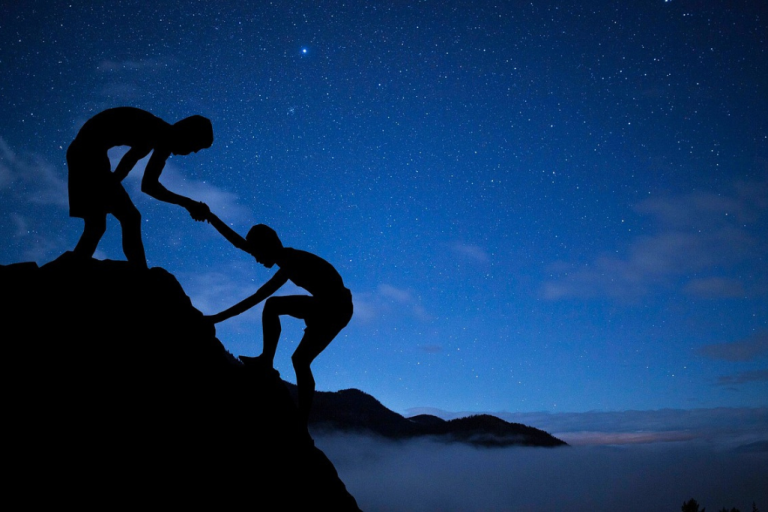 Two figures are silhouetted on a hillside against a dark blue night sky, one figure is bent over, helping the other figure to ascend the hill