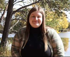 Maddie Baeten faces the camera, wearing a yellow/green plaid flannel over a black top. She has chest length brown hair.