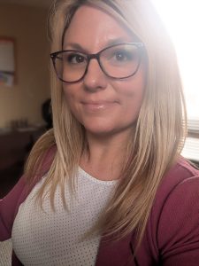 Jennifer wears a mauve cardigan over a white top. She has collar bone length blonde hair and dark large framed glasses.