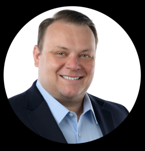 Dan is an adult man, facing the camera and smiling. He wears a blue collared shirt and suit jacket.