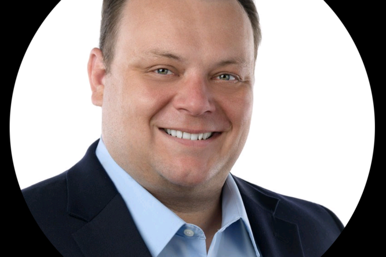 Dan is an adult man, facing the camera and smiling. He wears a blue collared shirt and suit jacket.