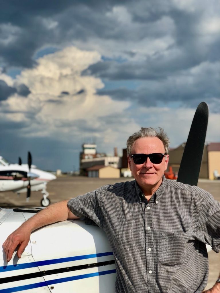 Fred Remer, smiling, at the front of his airplane with clouds in the background.