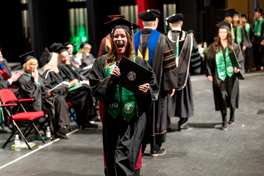 Person dressed in graduate regalia, crossing stage and holding diploma, mouth agape in excited smile