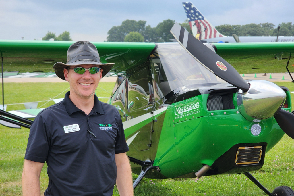 Man stands alongside a UND-green single-engine plane on an outdoor field.