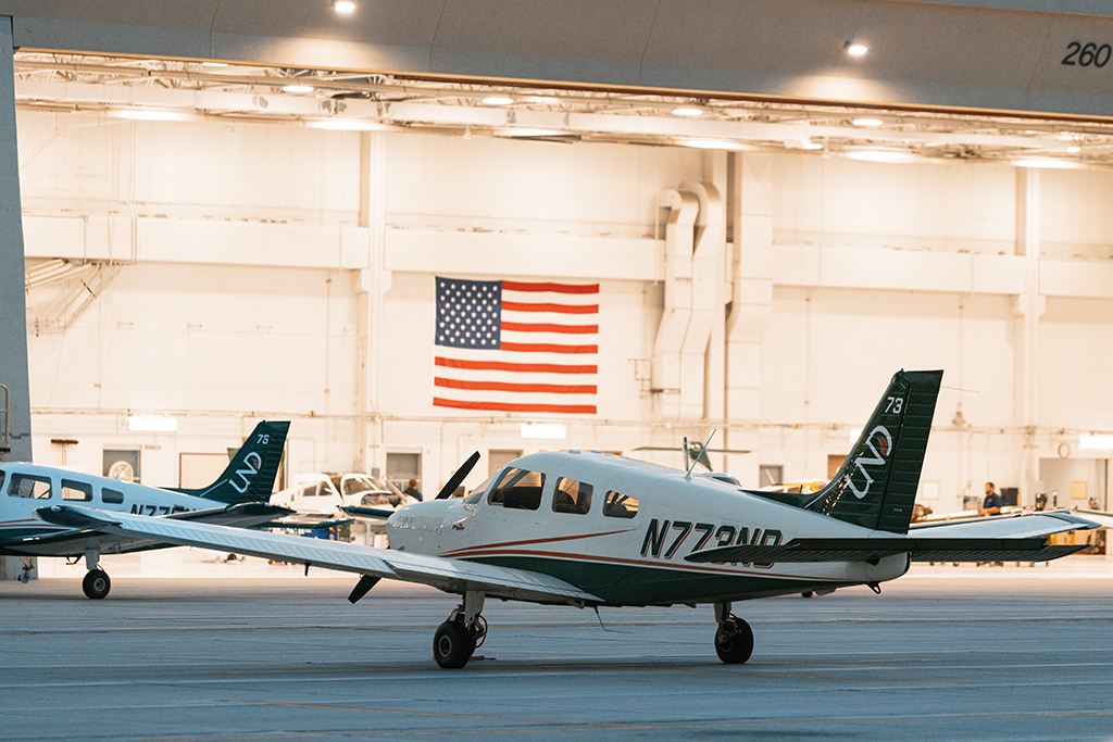 UND Aerospace aircraft sit on the ramp outside a brightly lit hangar, with an American flag hanging prominently on the back wall.