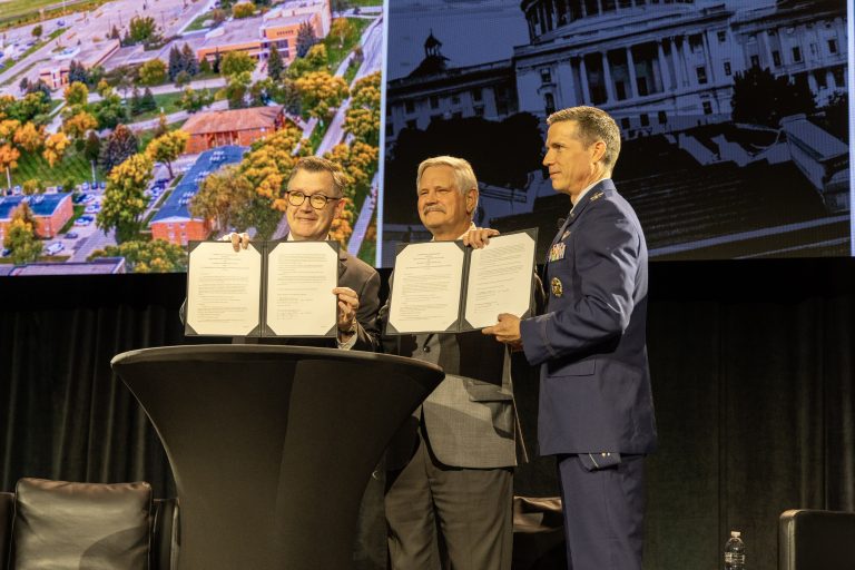 UND President Andrew Armacost, Sen. John Hoeven and Col. Jason Idleman stand onstage displaying copies of a signed memorandum of understanding.