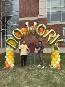 Three people standing under a balloon arch outside. 