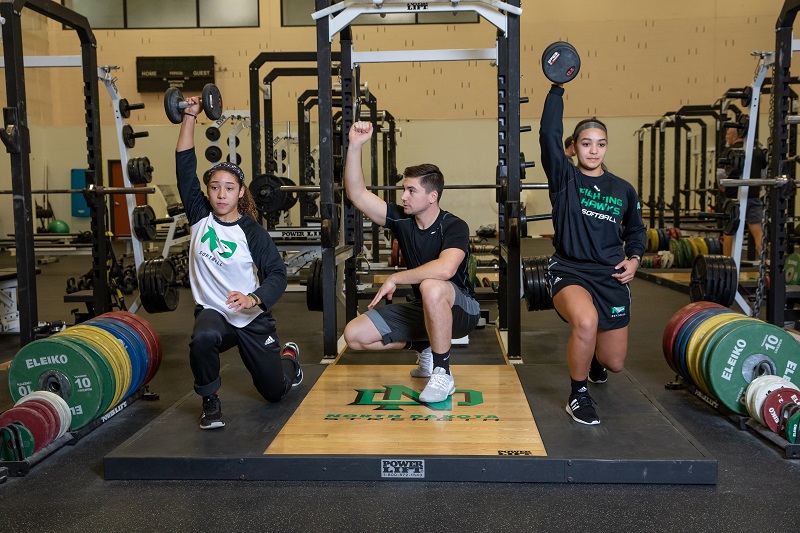 In this 2018 file photo, Trevor Dufner (center), a graduate student in kinesiology, works with UND student-athletes in the Hyslop Sports Center weight room. UND archival image.