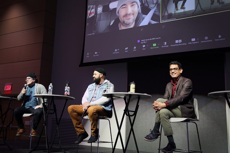 Three people sitting on stage as panelists with screen behind showing people participating virtually