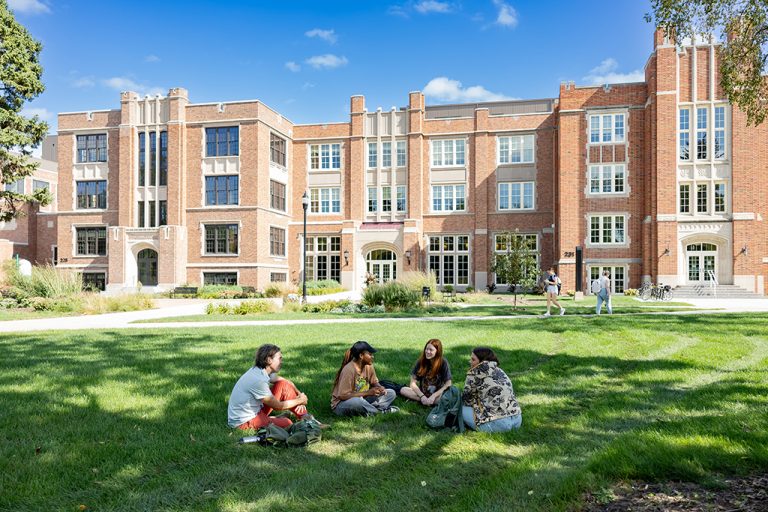 Students sitting together in grass of quad with campus buildings behind