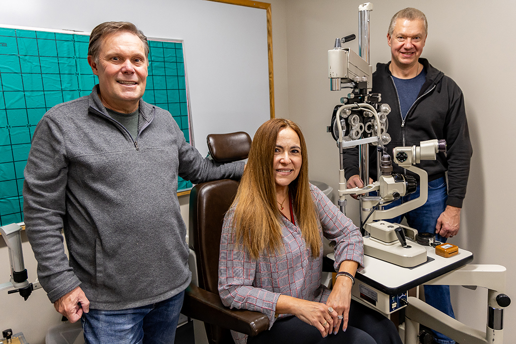 David Biberdorf, Carolina Praus Poltavski and Dmitri Poltavski pose for photo in eye examination room. Carolina sits in examination chair next to large set of eye examining machinery