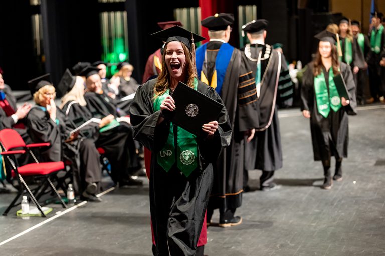 Person dressed in graduate regalia, crossing stage and holding diploma, mouth agape in excited smile