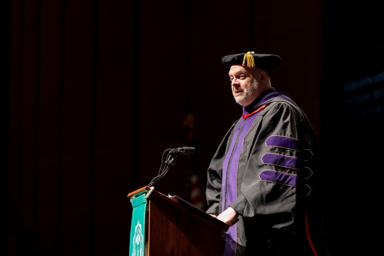 Dan Lewerenz at podium, speaking while wearing commencement regalia