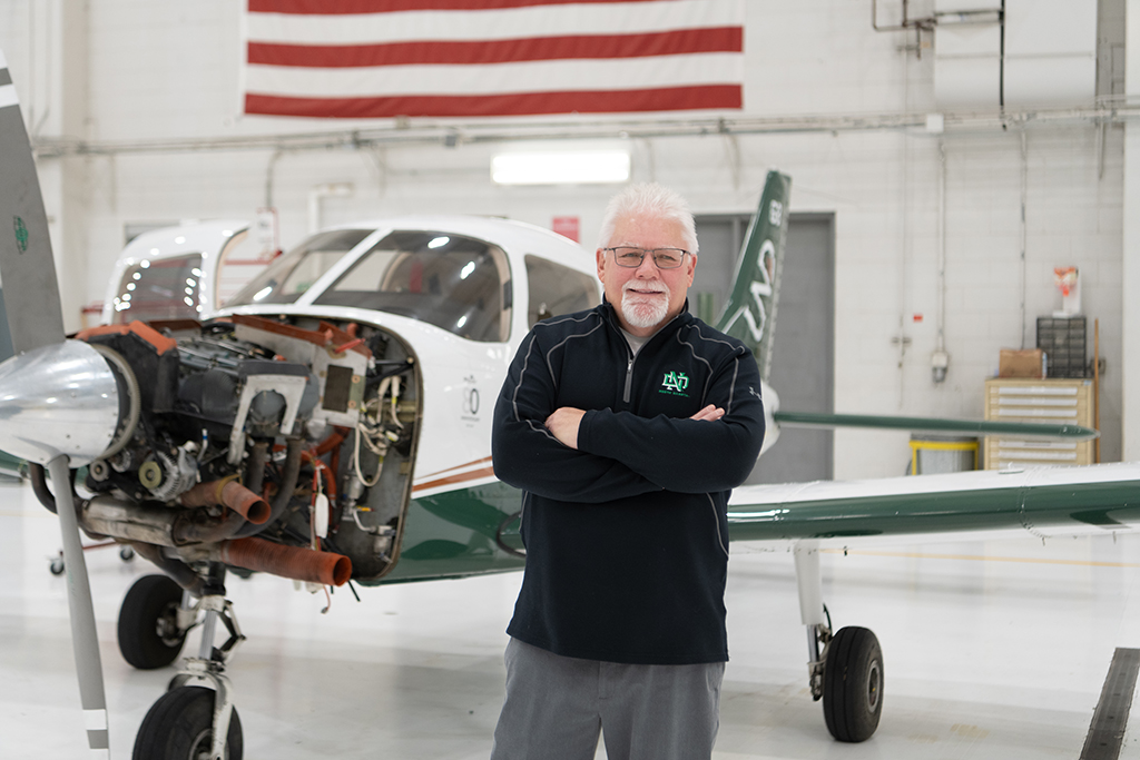 Dan Kasowski standing, arms crossed and smiling, in front of airplane in hangar. Airplane engine is exposed at nose of aircraft.