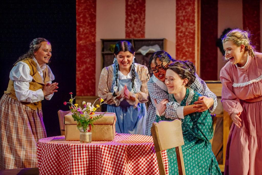 Five people dressed in costume, on set during theatre performance. One person embracing another seated at table as others look on
