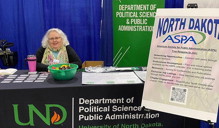 Michelle Buehlmann sitting at expo table for UND Department of Political Science & Public Administration near sign for North Dakota ASPA