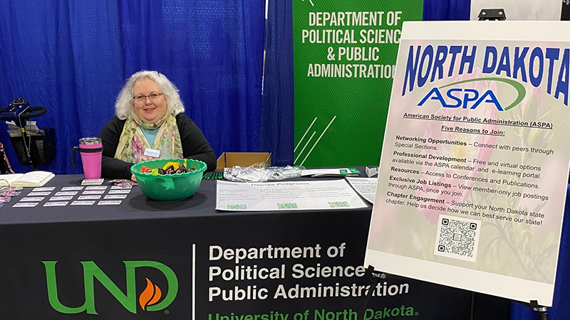 Michelle Buehlmann sitting at expo table for UND Department of Political Science & Public Administration near sign for North Dakota ASPA