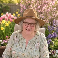 Deborah Radi sitting outside, wearing broad hat with blooming flowers in background