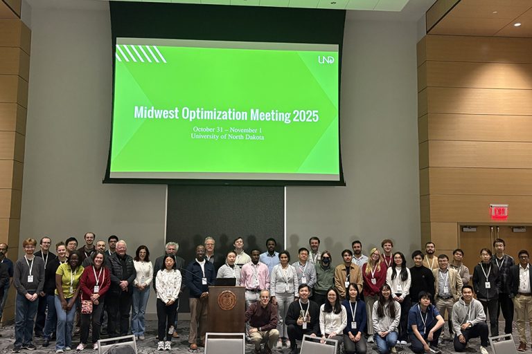Midwest Optimization Meeting attendees gathered for group photo under projection screen that reads "Midwest Optimization Meeting 2025"