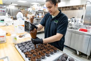 A student squeezes a generous swirl of chocolate frosting onto a batch of chocolate cupcakes. 