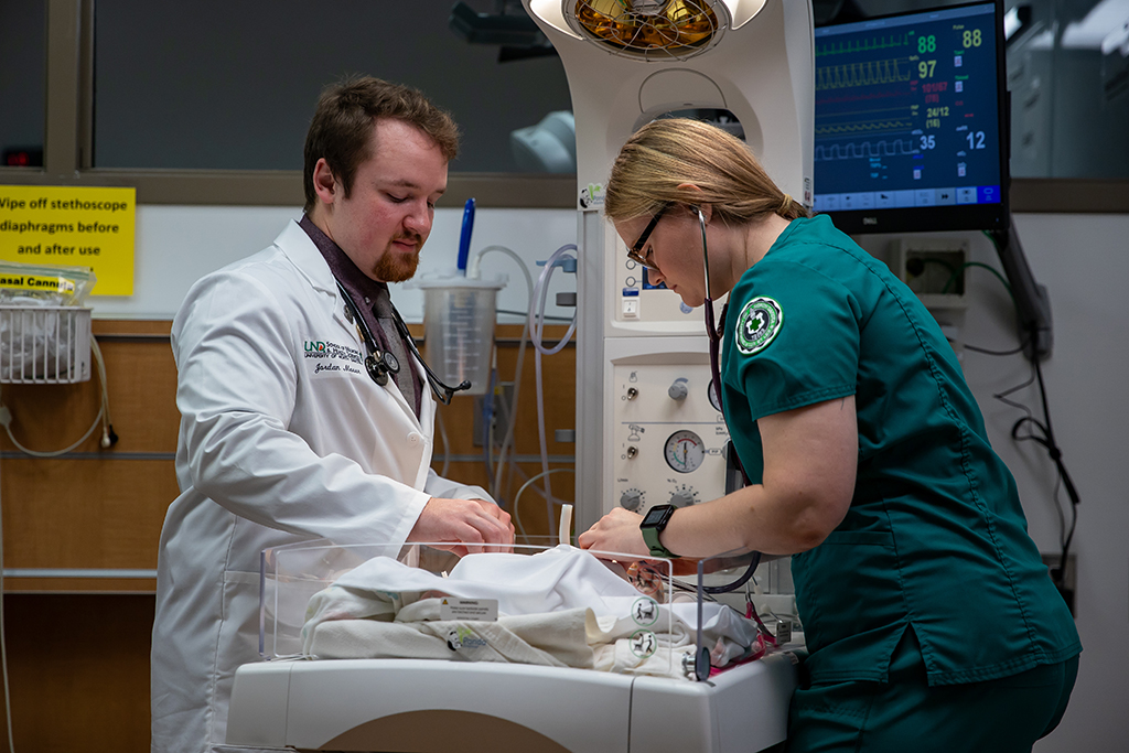 Two students, one wearing white coat and one wearing scrubs, work together over operating table.