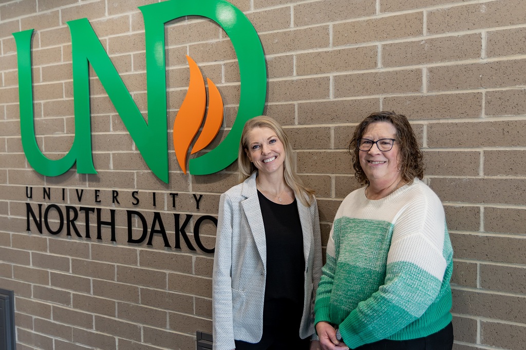 Two women stand by the UND logo in the entry way of the UND School of Medicine & Health Sciences.