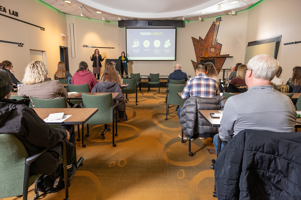A room full of people listen as two women talk near a podium up in the front.