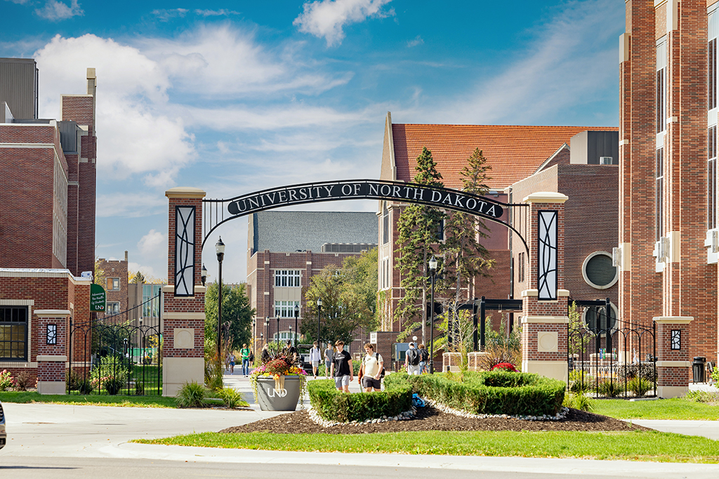 Archway on campus that says "University of North Dakota" across, students walk under with bright blue sky and campus buildings in background