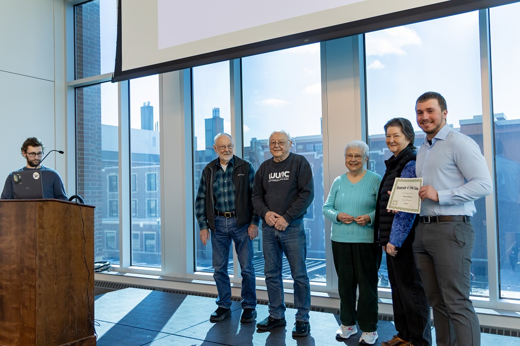 Student with certificate stands with other adults in front of a UND crowd