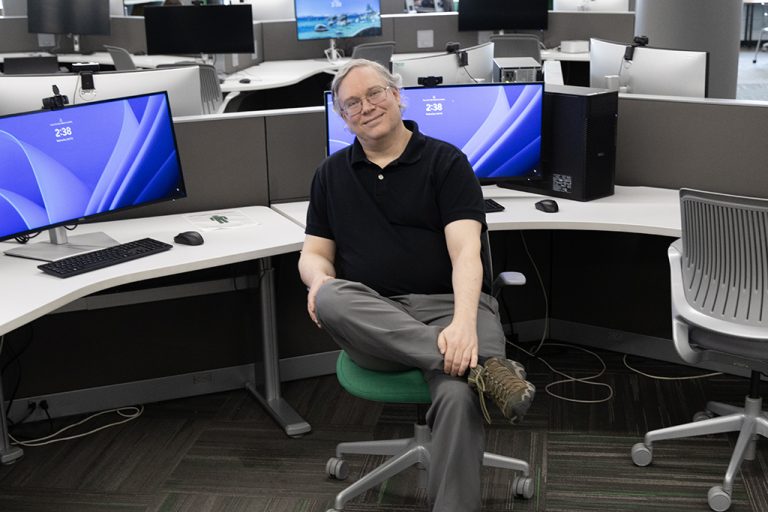 Will Martin sitting in chair, leg crossed, with rows of computer stations in background
