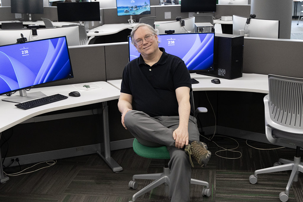 Will Martin sitting in chair, leg crossed, with rows of computer stations in background